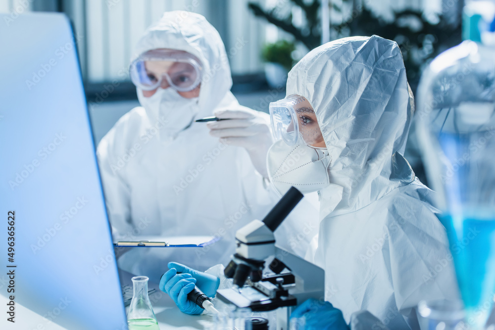 woman in hazmat suit working with microscope near computer monitor and blurred colleague. woman in hazmat suit working with microscope near computer monitor and blurred colleague.