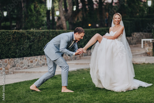 Картината върху платно Stylish groom in a gray checkered suit kisses the leg of the blonde bride in a long white dress in a meadow with green grass, against the background of the forest