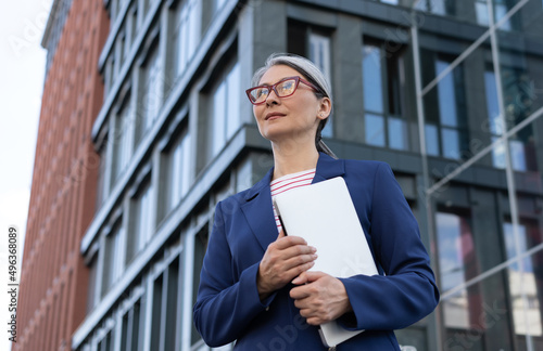 Confident mature businesswoman wearing red stylish eyeglasses holding laptop standing on urban street. Portrait of pensive asian manager waiting for taxi looking away outdoors. Successful business