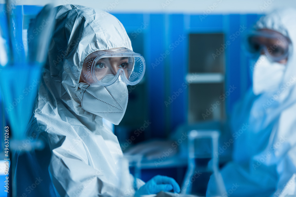 woman in goggles, medical mask and hazmat suit near scientist on blurred background. woman in goggles, medical mask and hazmat suit near scientist on blurred background.