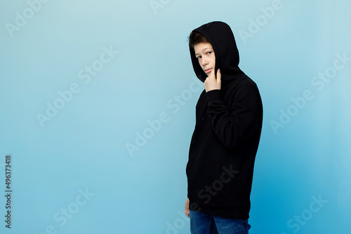 A thoughtful boy in a black hoodie and jeans looks at the camera on a monochrome blue background. There is a place for the inscription.