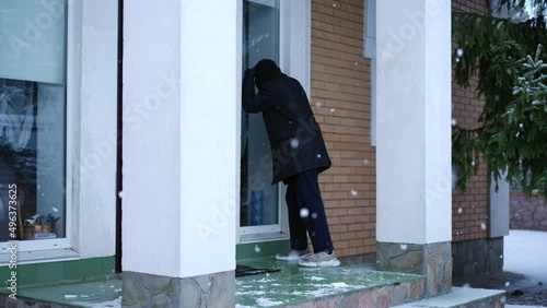 Wide shot male thief walking to house on snowy winter day looking inside. Caucasian man in balaclava ski mask examining empty home planning robbery. Crime and lifestyle photo