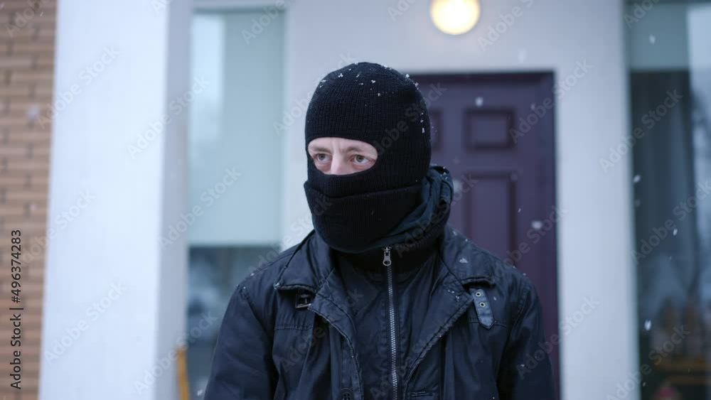 Portrait of suspicious Caucasian masked man looking around standing on snowy winter day outdoors. Young male robber at background of house planning burglary. Crime concept Portrait of suspicious Caucasian masked man looking around standing on snowy winter day outdoors. Young male robber at background of house planning burglary. Crime concept