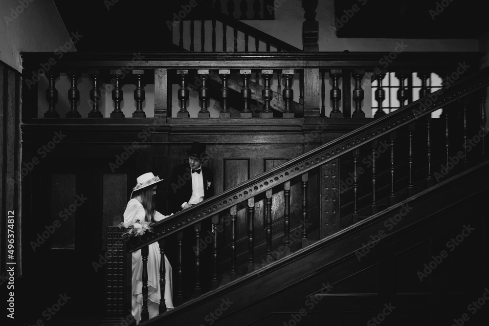Beautiful and elegant bride in a long blue dress and hat with bouquet in hands and her handsome groom in a suit are standing in the dark room with sunlight. Beautiful and elegant bride in a long blue dress and hat with bouquet in hands and her handsome groom in a suit are standing in the dark room with sunlight.