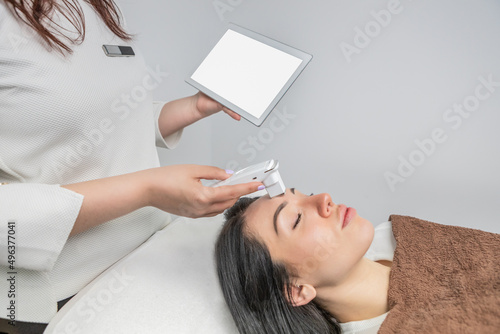 Woman receiving a facial analysis and diagnosis. Dermatologist holds a tablet to find out the state of the skin. Detailed scan of skin hydration, sebum, acne, wrinkles, sensitivity and melanin. 