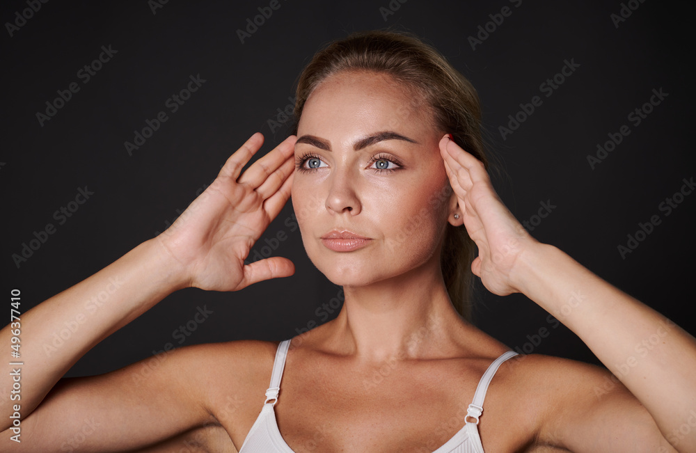 Close-up portrait of an European pretty woman with fresh glowing skin holding hands on her temples, doing massage movements on her face. Anti-aging concept, smoothing, rejuvenating beauty treatment
