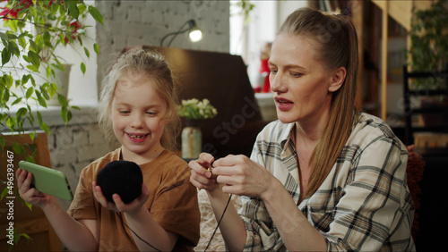 A girl helps her mother to knit. She holds a cell phone in one hand and a ball of nits in the other