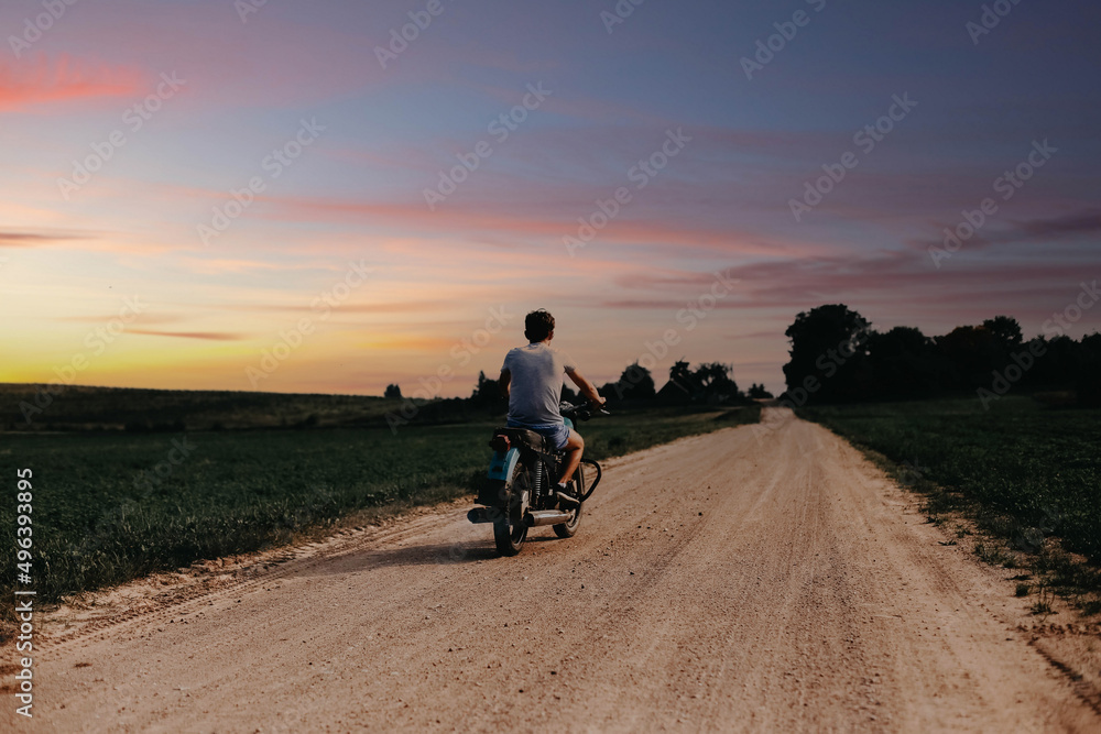 young man rides on a country road on a retro vintage motorcycle, a biker rides at sunset.