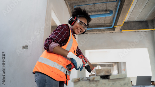 Cute looking female engineer with afro hairstyle. African descent  cordless electric drill screws to planks at a construction site.She wears safety glasses Anti-noise earmuffs,gloves,reflective vest.