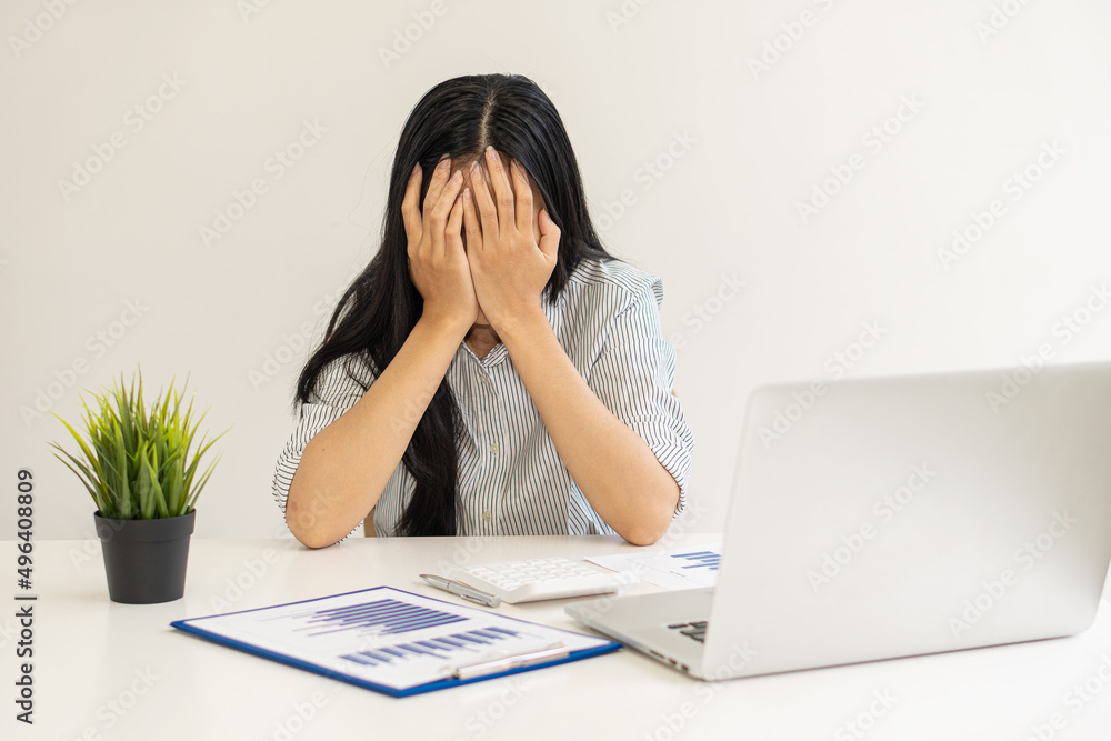 A young Asian businesswoman wearing a blue shirt covering her face with her hands and feeling stressed working with a laptop computer and graph paper on a desk in a financial accounting concept office A young Asian businesswoman wearing a blue shirt covering her face with her hands and feeling stressed working with a laptop computer and graph paper on a desk in a financial accounting concept office