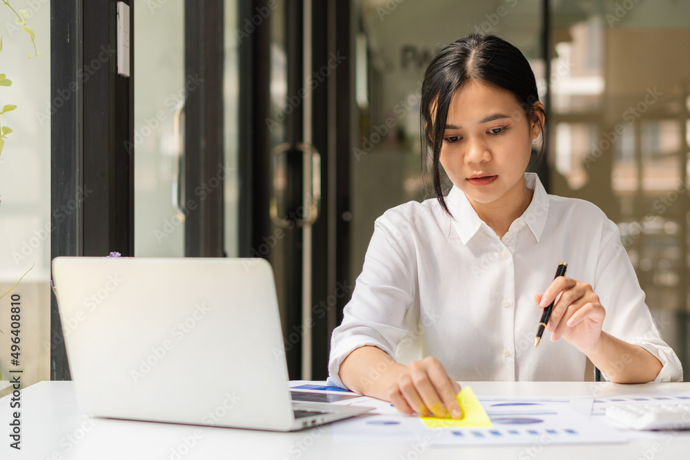 Asian female accountant or banker calculating savings, finance, and economy concepts via laptop. Businesswoman writing charts and graphs that show results in computational analysis. Asian female accountant or banker calculating savings, finance, and economy concepts via laptop. Businesswoman writing charts and graphs that show results in computational analysis.