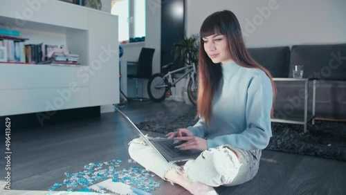 Young woman sitting cross legged at home typing on laptop smart device sitting cross legged in her modern living room with puzzle pieces all around the wooden parquet floor wearing ripped jeans photo