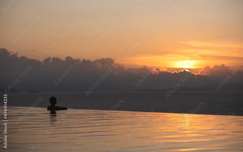 A young woman relaxing in infinity swimming pool and looking at a beautiful sunset and the sea view