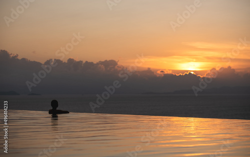 A young woman relaxing in infinity swimming pool and looking at a beautiful sunset and the sea view © Farknot Architect