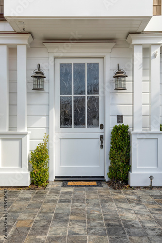 Front door that is white in color with two lamp fixtures  potted plants and a cool tile pattern showing off the entrance.