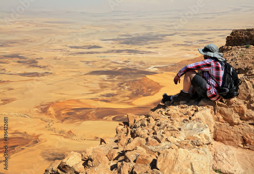 Traveler with a backpack sitting on the cliff. Man looks into the distance at color sand stone Negev desert. National geological erosion land form park HaMakhtesh HaGadol. Large Crater. Israel