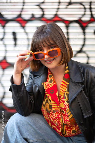 Young caucasian woman posing with style next to a graffiti wall. She wears a cute trendy outfit and sunglasses. Female outdoor portrait.