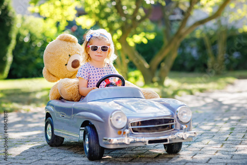 Little adorable toddler girl driving big vintage toy car and having fun with playing with plush toy bear  outdoors. Gorgeous happy healthy child enjoying warm summer day. Smiling stunning kid in gaden