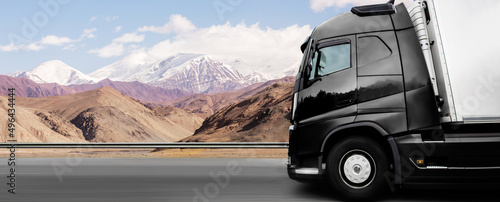 Side view of a bright black large semi-trailer truck carrying cargo in a long semi-trailer on a flat road with a mountain landscape and clouds in the background