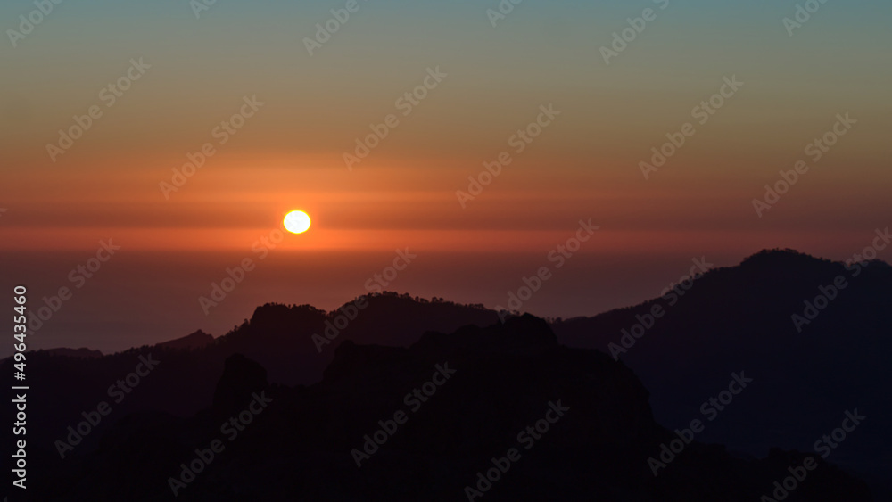 Stunning sunset above the rugged mountains of island Gran Canaria, Canary Islands, Spain with red sun in the colorful sky and the silhouettes of hills. Stunning sunset above the rugged mountains of island Gran Canaria, Canary Islands, Spain with red sun in the colorful sky and the silhouettes of hills.