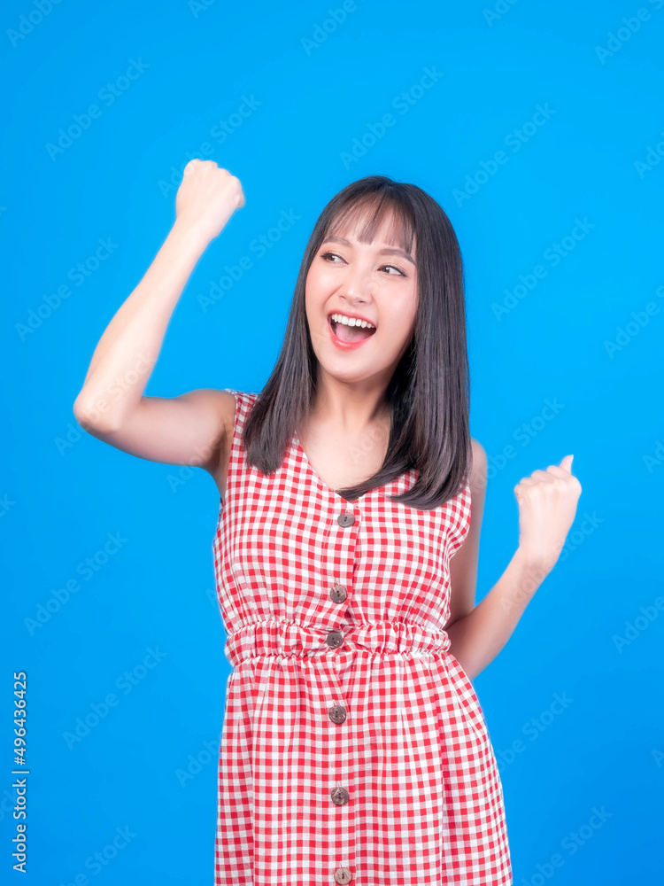 Fototapeta premium Portrait of excited Beautiful Asian woman cute girl in red dress with bangs hair style raising hand in the air laughing cheerfully gesturing for winner , smiling , look camera isolated blue background