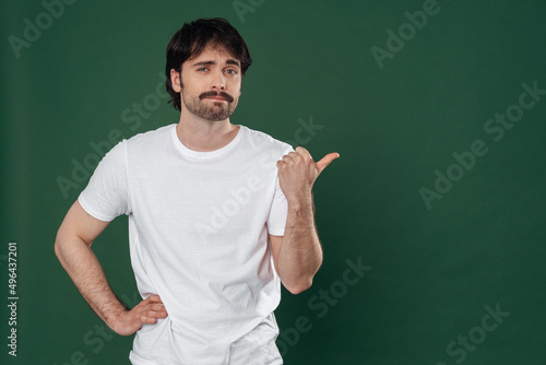 On a dark green background, a young man in a white T-shirt points his thumb to the right © Оlgart
