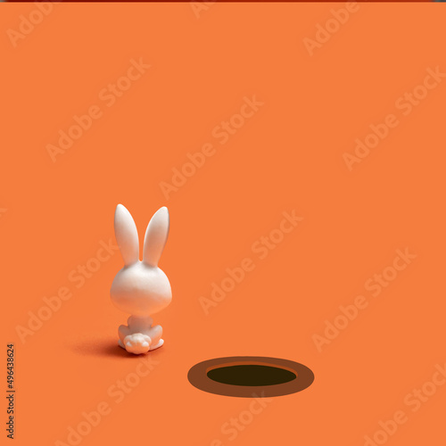 Down to the rabbit hole minimal concept. Cute little white rabbit toy calmly sits near a dark hole. Creative copy space above against terracotta background.