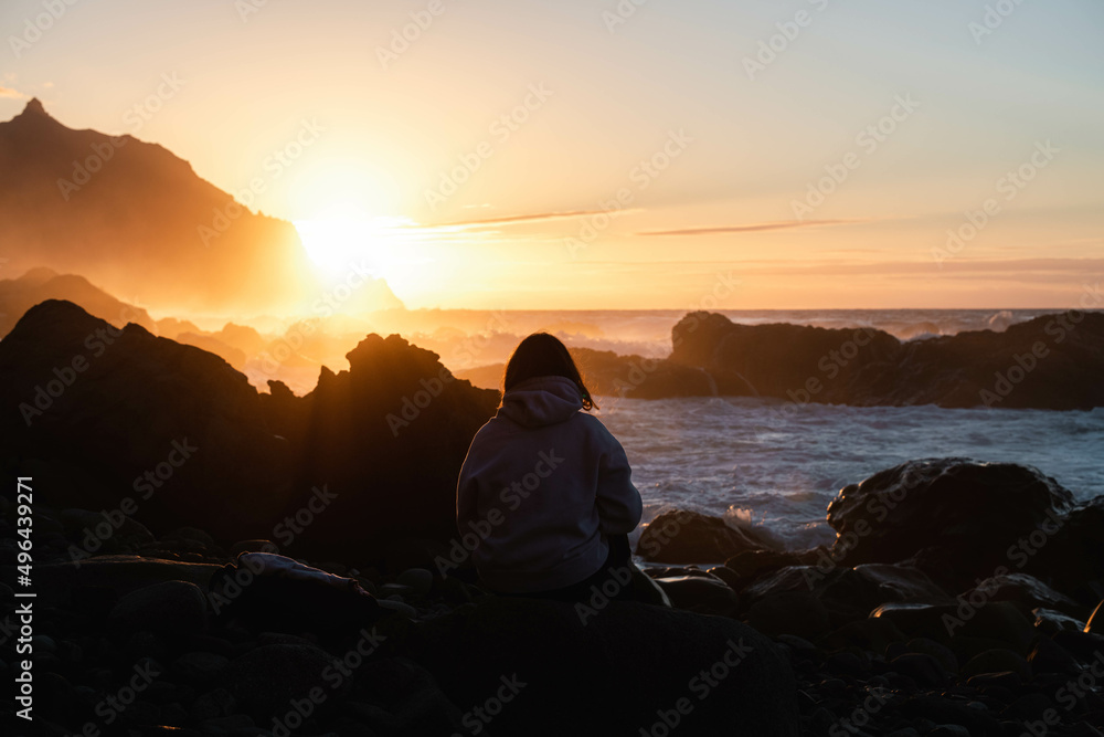 Brown haired woman seen from behind watching the sunset sitting on the beach with the waves crashing in the background