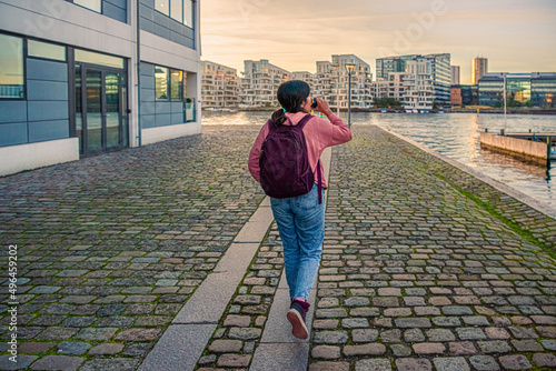 Photography A young girl walks along a pedestrian road with paving stones on the embankment along the water canal and drinks against the backdrop of residential buildings