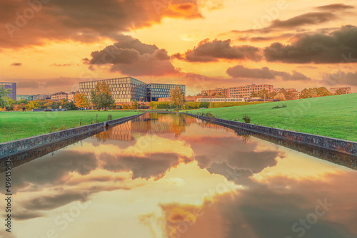 Fotografie Beautiful yellow sunset with clouds over a river channel with reflection in a park overlooking residential buildings and a metro station DR Byen in the city area Ørestad