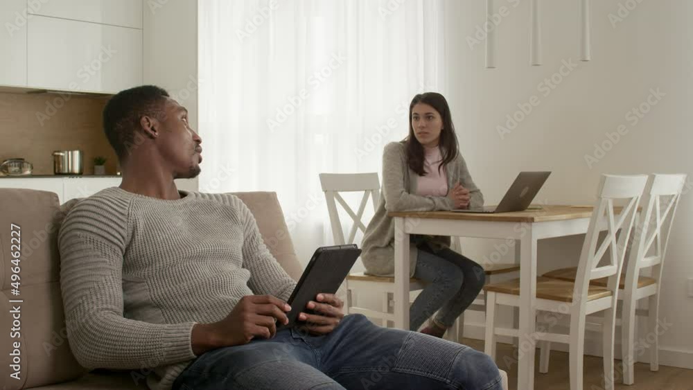 Mixed race couple chilling at home, browsing the web on a tablet and working on a laptop at home Mixed race couple chilling at home, browsing the web on a tablet and working on a laptop at home