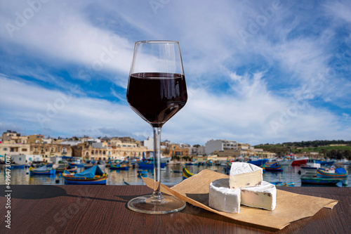 Glass of red wine with brie cheese with view of harbor with traditional boats and historic city center in Marsaxlokk, Malta photo