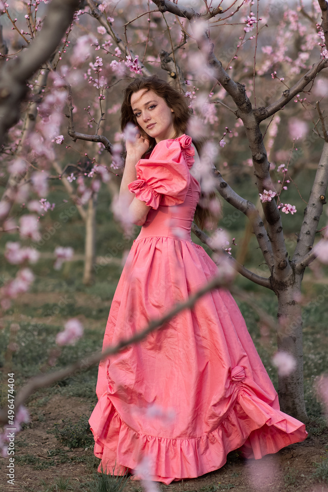 Flowering trees in spring. A beautiful young European girl in a pink dress stands in gardens with peach trees. Flowering trees in spring. A beautiful young European girl in a pink dress stands in gardens with peach trees.