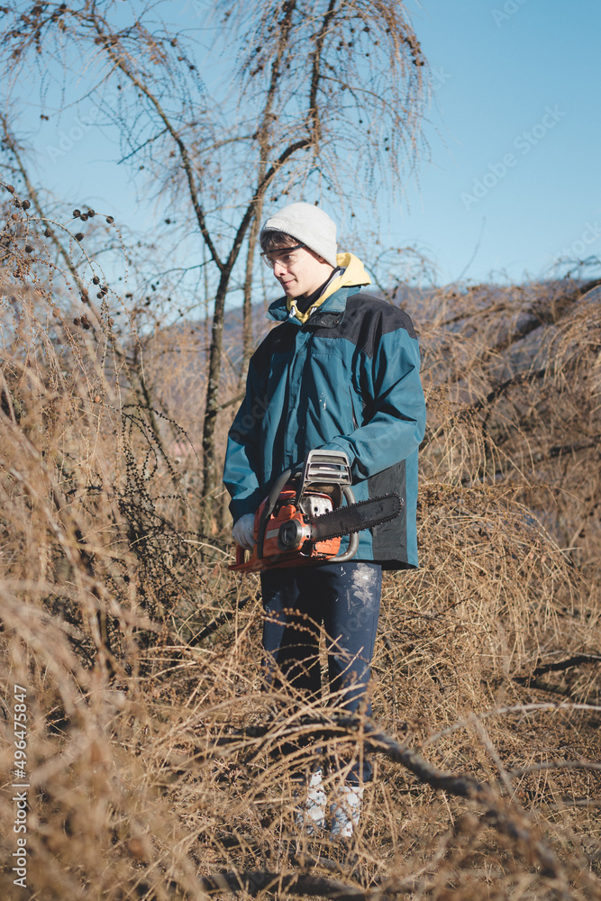 young 17-year-old temporary worker in work clothes wrestles with a chainsaw and a dry larch tree. Wood processing in the wilderness. Cutting a fallen tree into parts. Learning a new workforce young 17-year-old temporary worker in work clothes wrestles with a chainsaw and a dry larch tree. Wood processing in the wilderness. Cutting a fallen tree into parts. Learning a new workforce