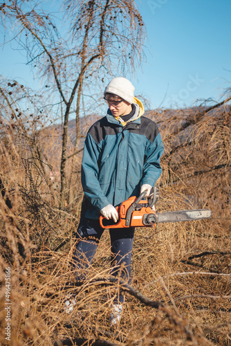 young 17-year-old temporary worker in work clothes wrestles with a chainsaw and a dry larch tree. Wood processing in the wilderness. Cutting a fallen tree into parts. Learning a new workforce