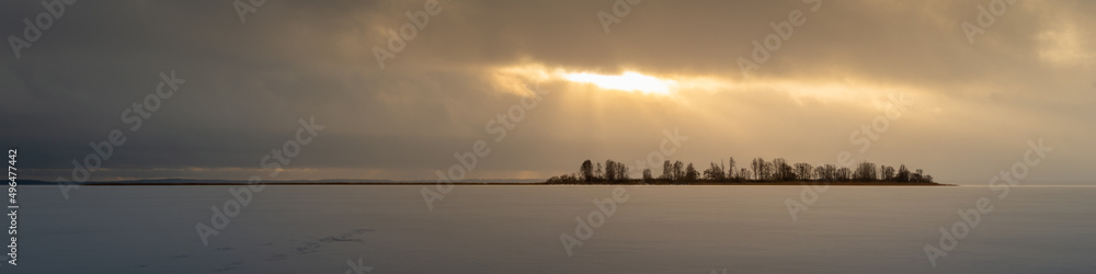 dramatic winter landscape. wide panoramic view of a frozen snow-covered lake with an island lit by sunbeams through a gap among a cloudy sky