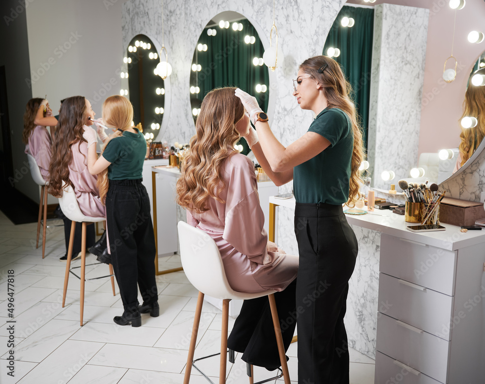 Beauty specialists doing professional makeup for clients in modern salon. Young woman with long curly hair sitting at dressing table while female stylist applying foundation with sponge.