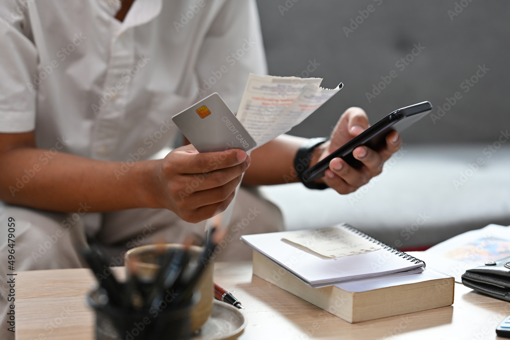 Fototapeta premium Close up with man sitting on a couch while holding bills and credit card and using mobile phone for calculate, finance, business, home and technology concept.