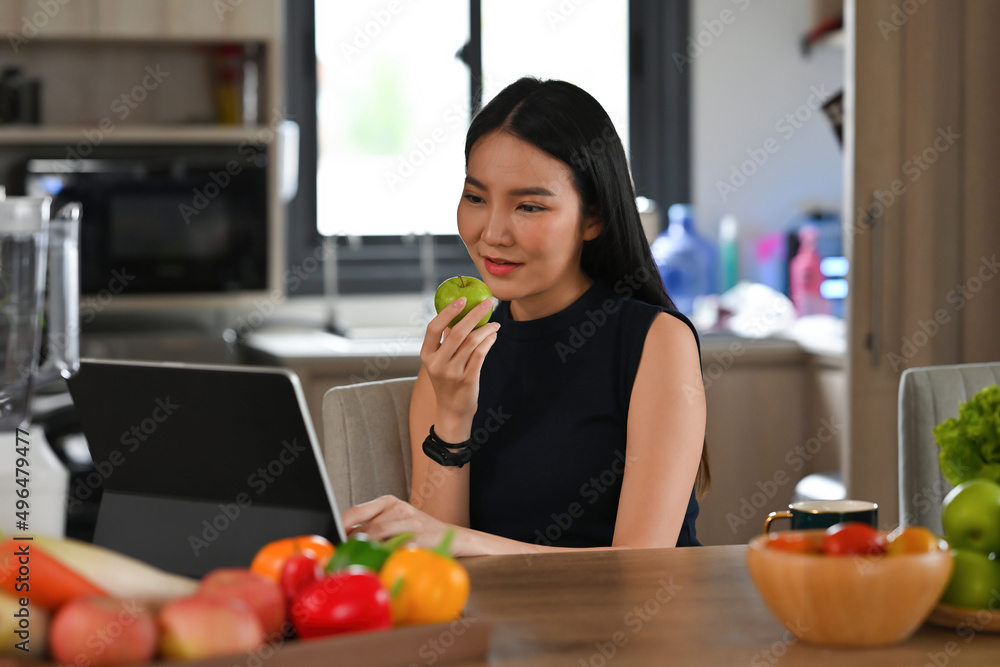 A portrait of a young pretty Asian woman chopping fruits and preparing a meal on a wooden cut board, having a tablet to look at a recipe, for food, health and cooking concept.
