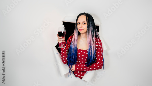 Young woman with glass of wine in studio. Beautiful female in red dress drinking alcoholic beverage in hole of white background.