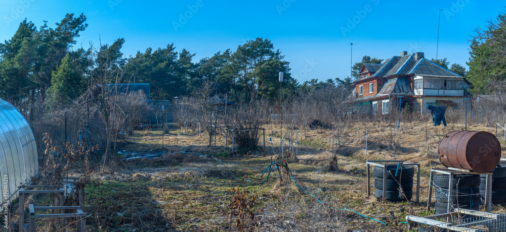 An unkempt plot of land in the village in early spring with withered grass, a greenhouse and a rusty barrel.