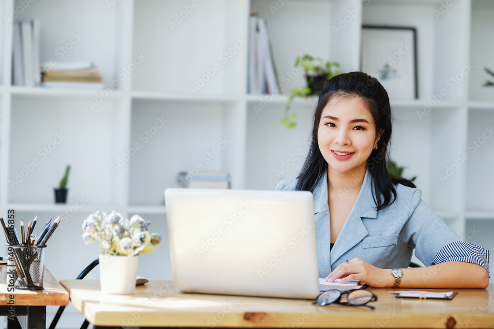 An Asian entrepreneur or businesswoman shows a smiling face while working with using computer on a wooden table. An Asian entrepreneur or businesswoman shows a smiling face while working with using computer on a wooden table.