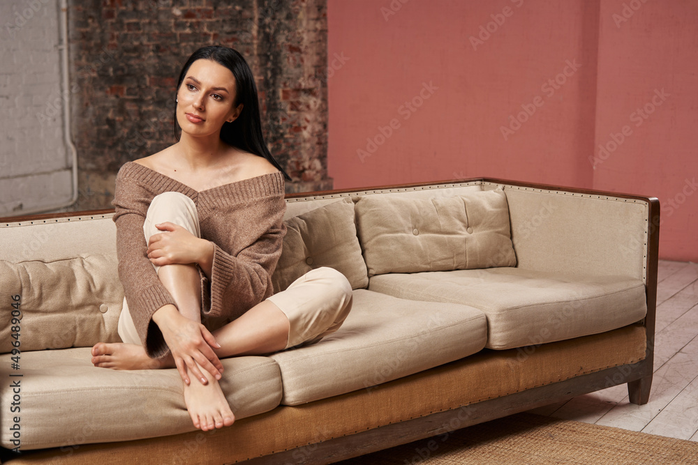 a beautiful woman with black hair in cozy clothes of a natural shade sits on a sofa with her foot on the sofa and holds on to it with her hands. cozy time at home. calmness and comfort a beautiful woman with black hair in cozy clothes of a natural shade sits on a sofa with her foot on the sofa and holds on to it with her hands. cozy time at home. calmness and comfort
