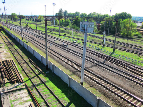 Evenly laid railroad rails crossing the frame diagonally. Railroad track, viewing platforms, rails for repairs, behind the fence. Wires next to greenery. Transfer switches, traffic lights and mechanis
