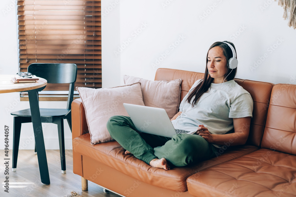 Obraz premium Modern professional woman working in headphones on a laptop while sitting on the couch at home.