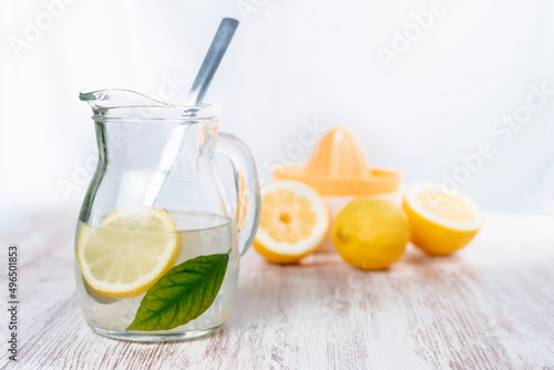 Pitcher with lemonade and cubes, a slice of lemon and a green leaf, in the background some lemons and a squeezer out of focus.