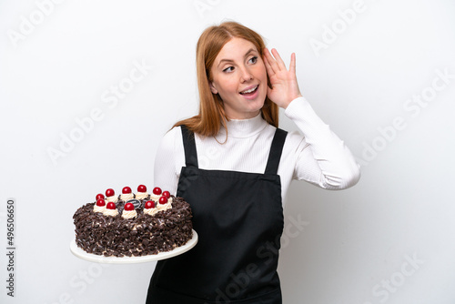 Young redhead woman holding birthday cake isolated on white background listening to something by putting hand on the ear