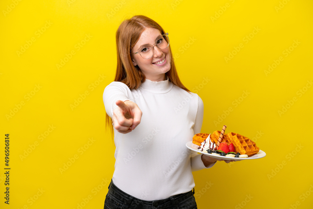 Young redhead woman holding waffles isolated on yellow background pointing front with happy expression Young redhead woman holding waffles isolated on yellow background pointing front with happy expression
