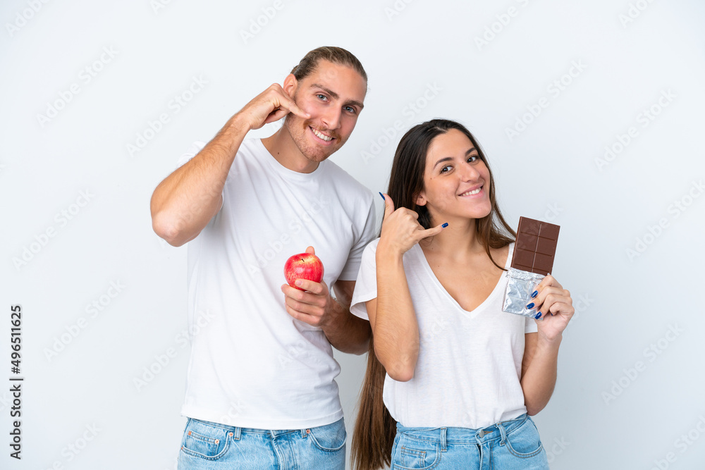 Young caucasian couple with chocolat and apple isolated on white background making phone gesture. Call me back sign Young caucasian couple with chocolat and apple isolated on white background making phone gesture. Call me back sign