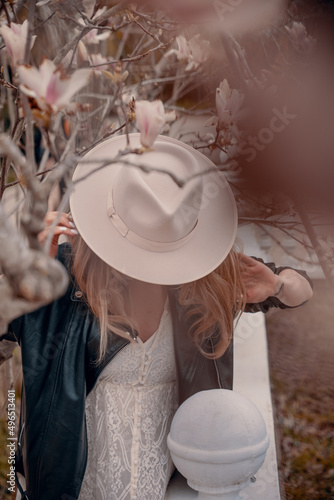 Beautiful young woman dressed  vintage jacket and white hat, standing in magnolia tree, with big  pink magnolia flowers. Shallow focus and depth of field